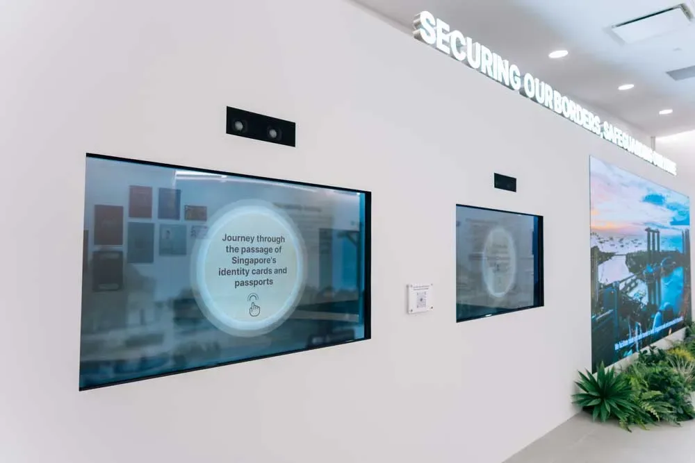 Two touchscreens set into a white wall. The nearest one invites visitors to journey through the passage of Singapore's identity cards and passports