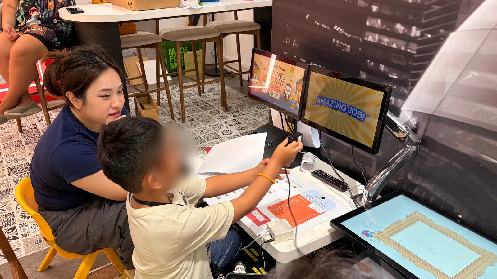 A child holding a card up to a reader at a kiosk with two screens, one showing Amazing job!, with a researcher beside them. The child's face is blurred.