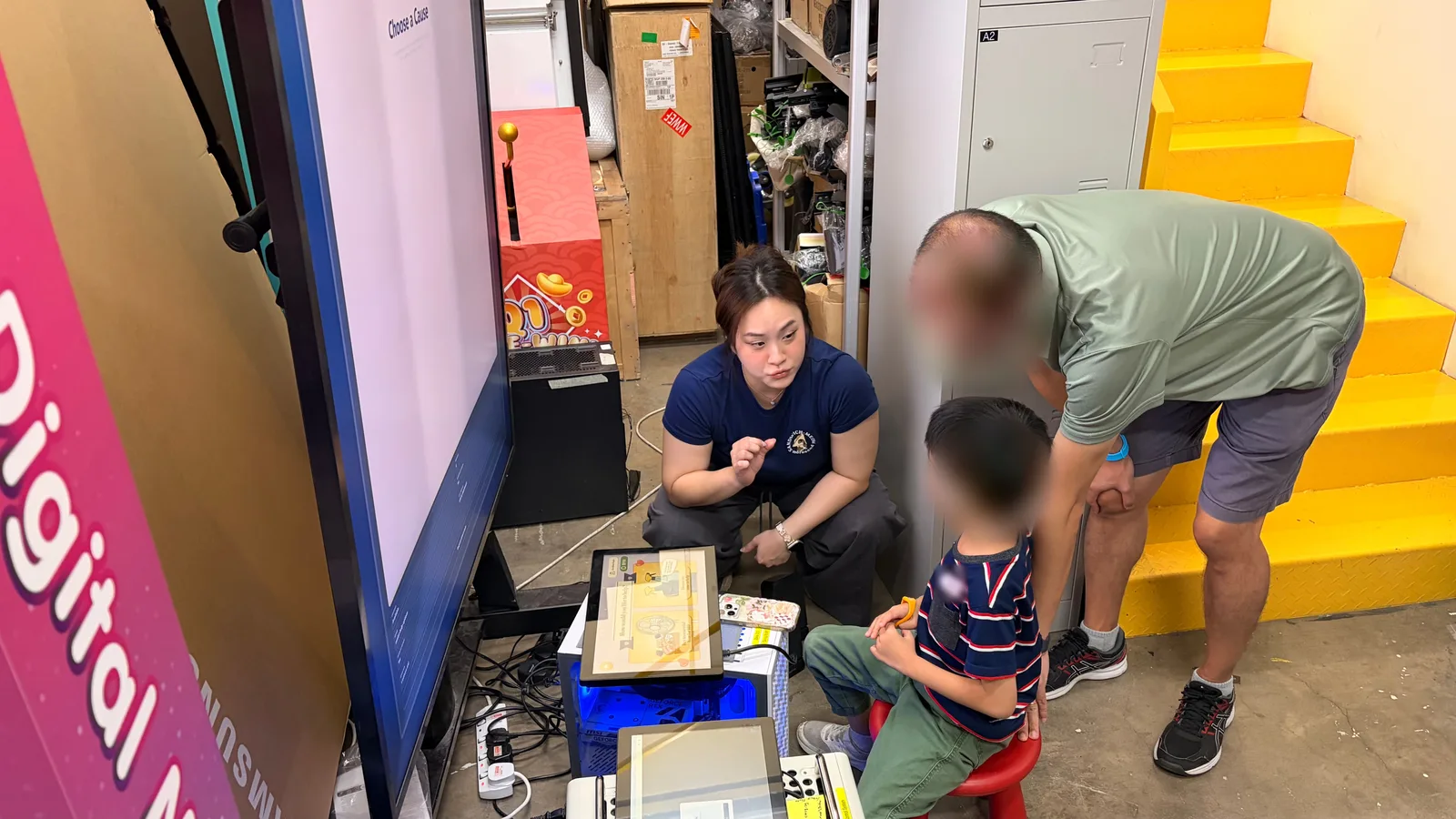 A researcher crouching beside two kiosk touchscreens to talk with a seated child, while a parent leans in. The child's and parent's faces are blurred.