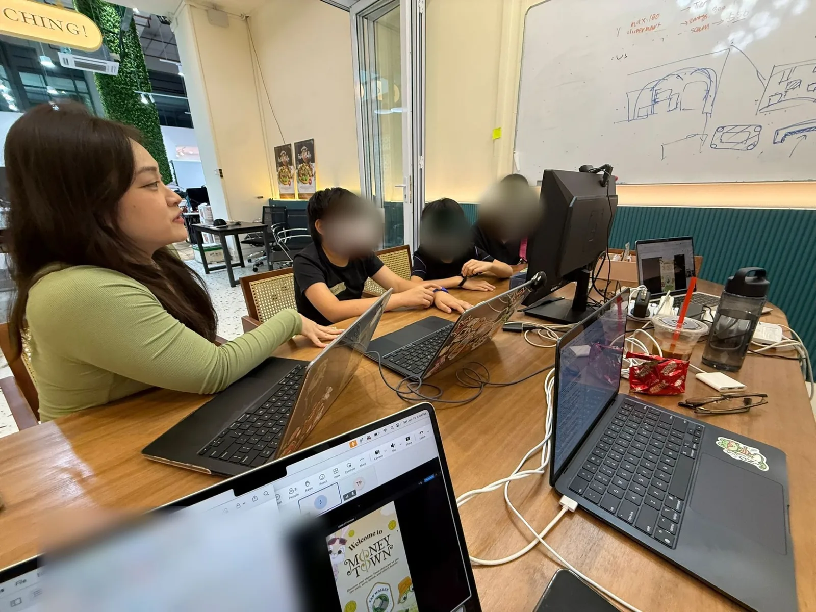 A testing session at a long wooden table, with a researcher and two children at laptops and a parent beside them, and a whiteboard of sketches behind. The children's and the parent's faces are blurred.